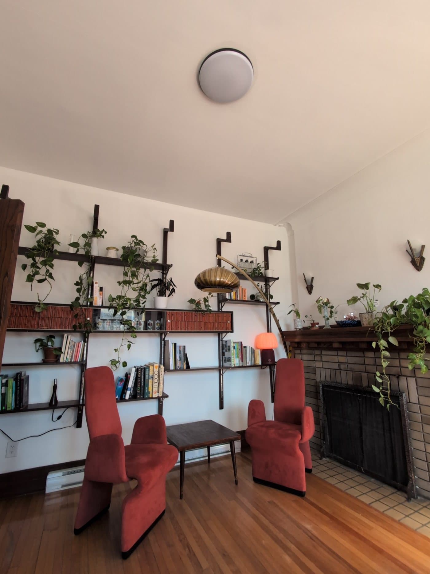 Salon du bureau d'Atelier FlorA : bibliothèque murale remplie de plantes et de livres, deux fauteuils rouges, une lampe arquée et un foyer en brique