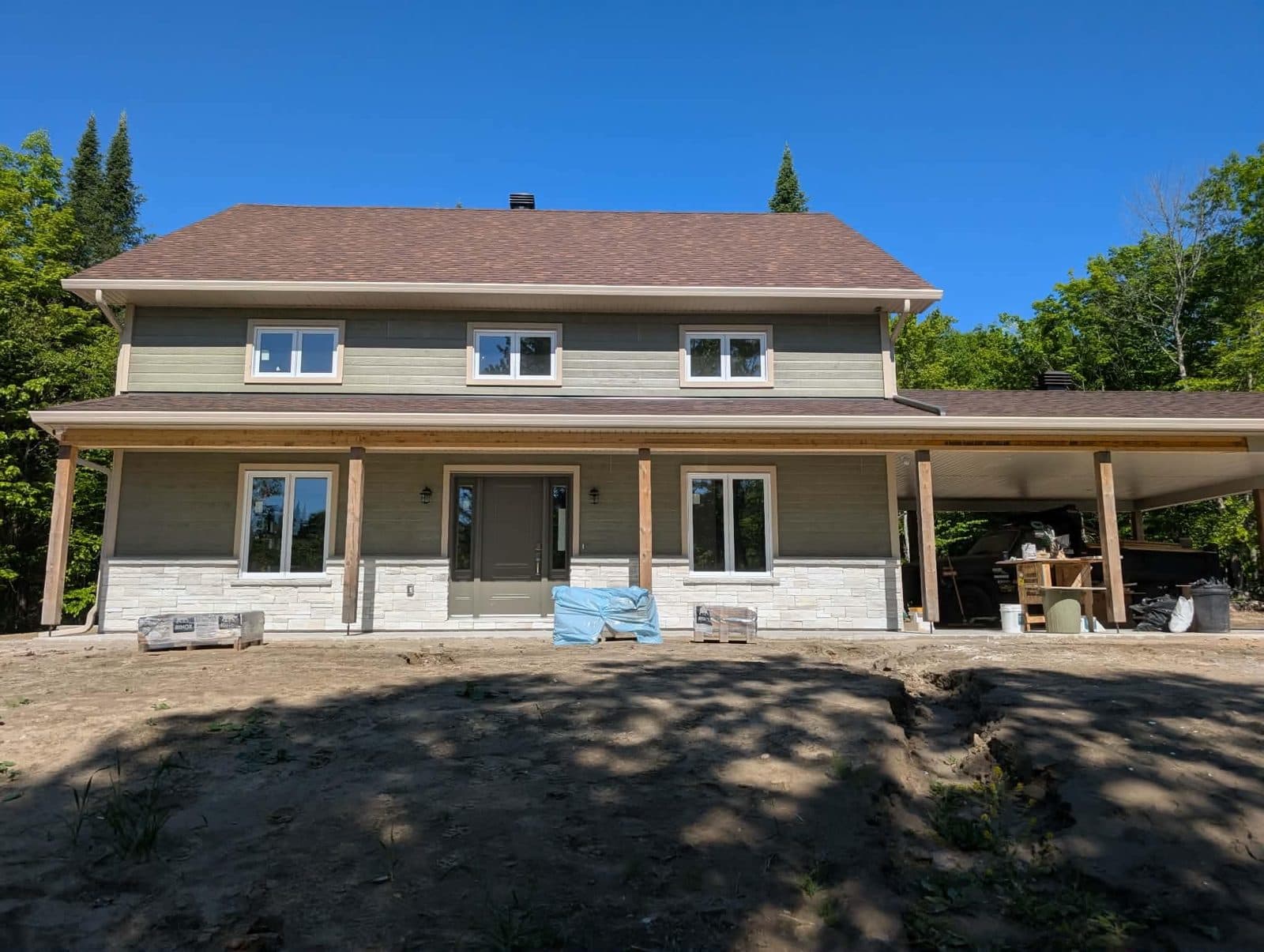 Maison neuve à étage au revêtement gris, à la base de pierre et à la galerie couverte, photographiée en fin de chantier