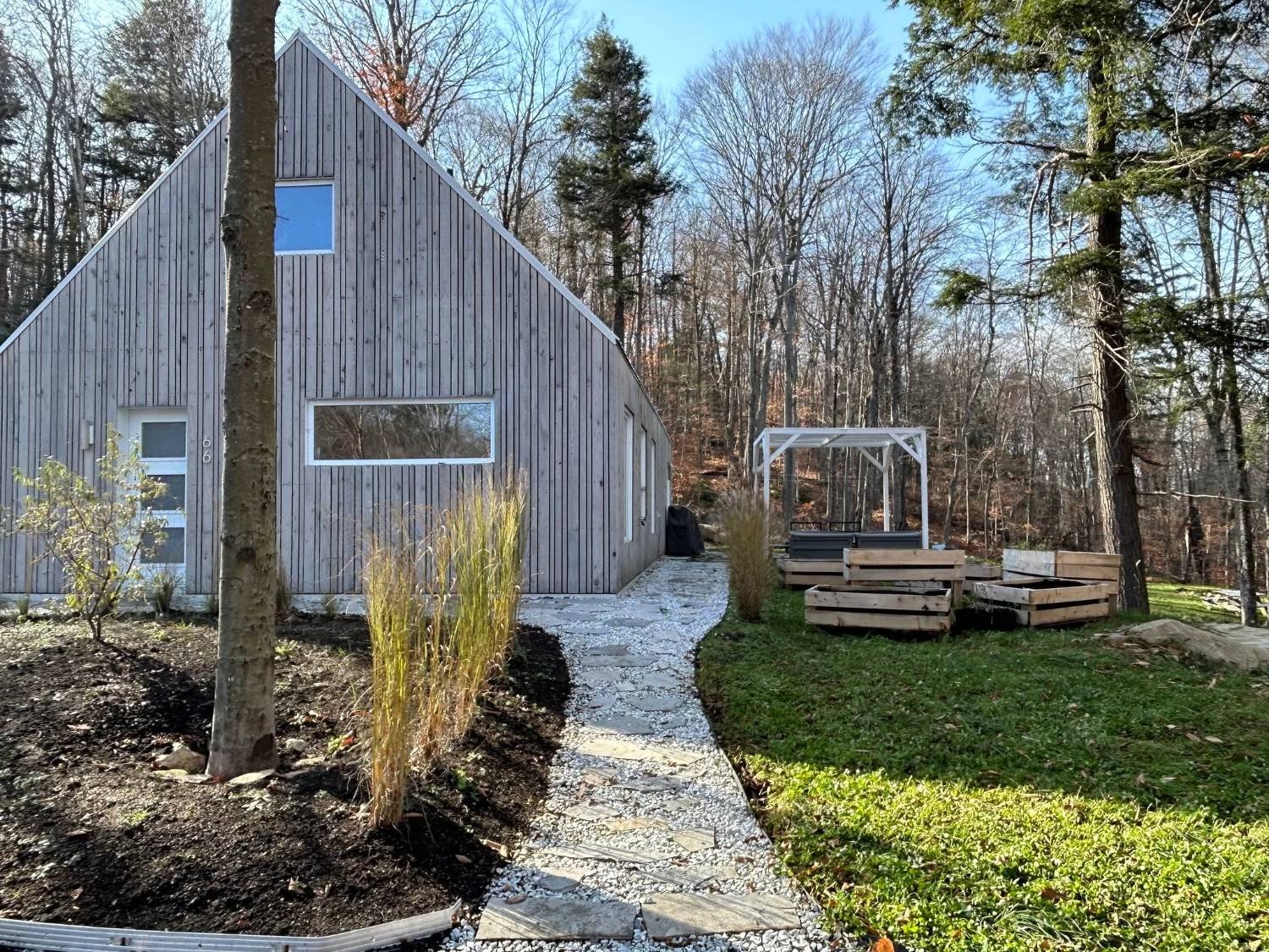 Résidence en forêt vue de côté : bardage de bois, sentier de pierre, pergola blanche et bacs de potager